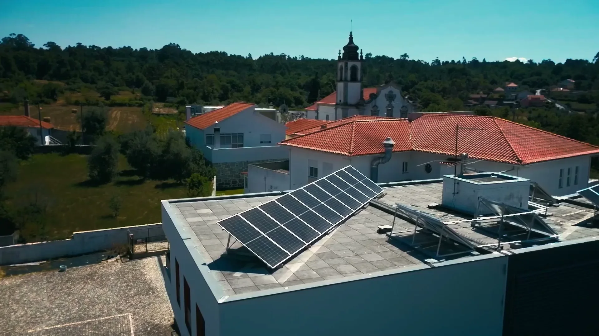 Vista aérea da cobertura do Centro Social da Paróquia de Boa Aldeia, com os painéis fotovoltaicos e os coletores solares térmicos instalados e a igreja da paróquia ao fundo.
